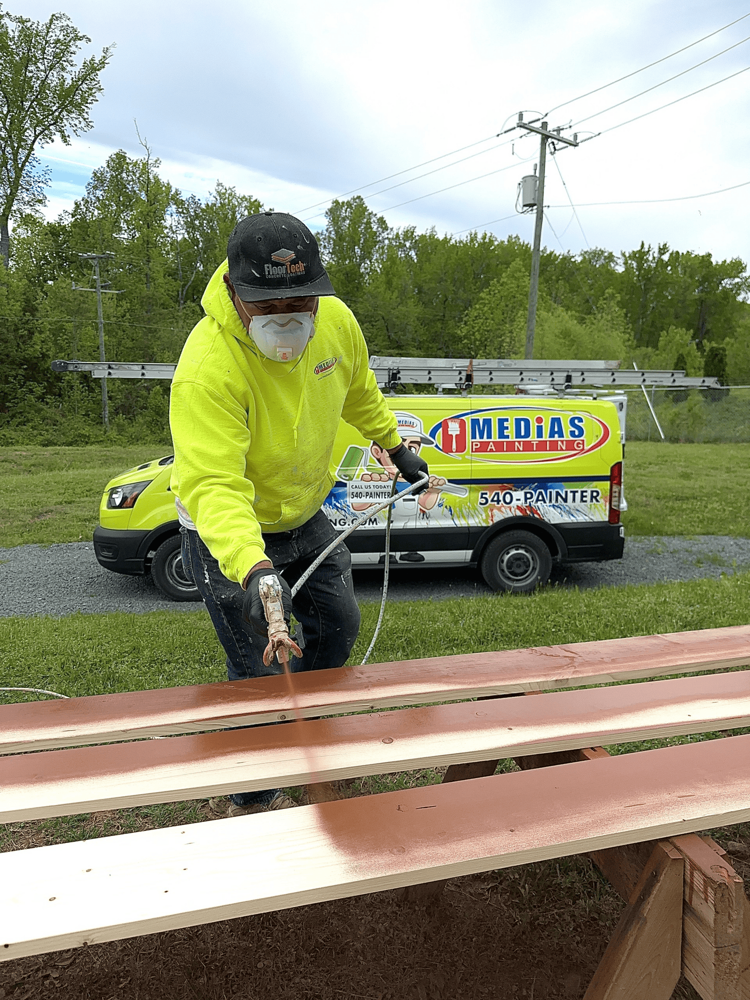 Professional Deck Staining and Wood Finishing Services A professional painter wearing a mask spray staining wooden deck boards outdoors with a painting company van.