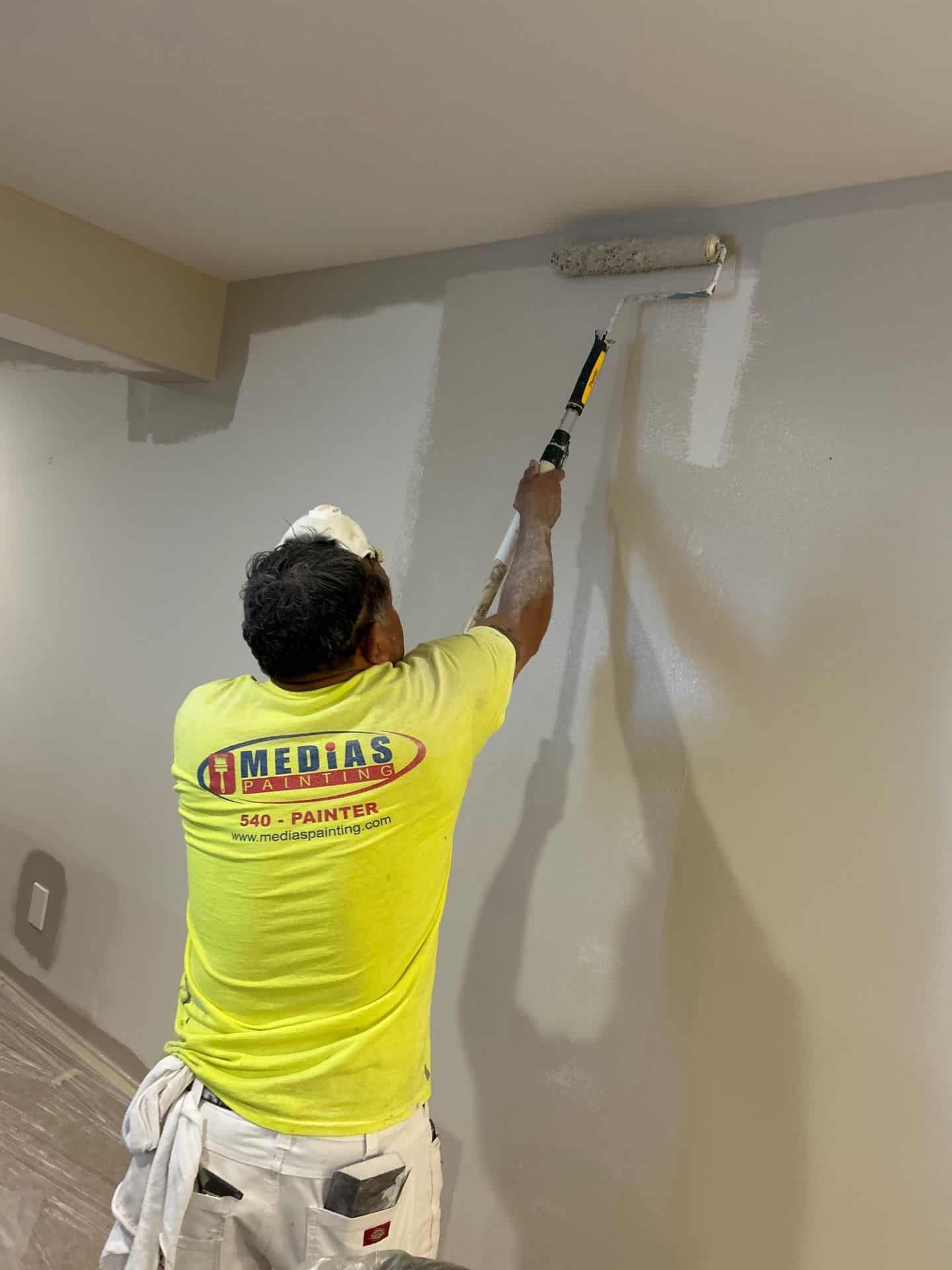 A painter from Medias Painting applies light gray paint to an interior wall using a roller.