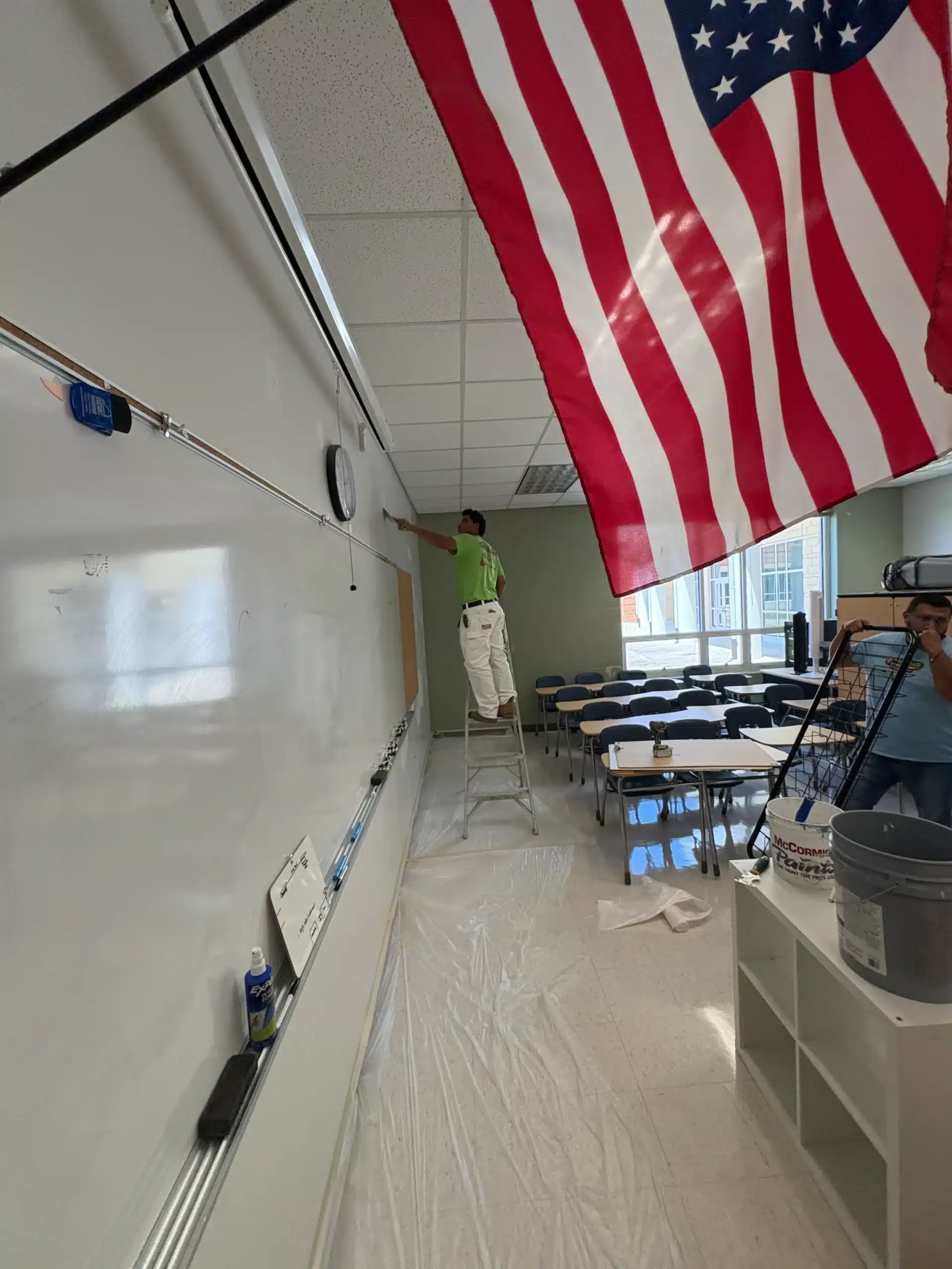 Classroom Interior Painting Project Painter in lime-green shirt on ladder painting white classroom wall with American flag hanging from ceiling